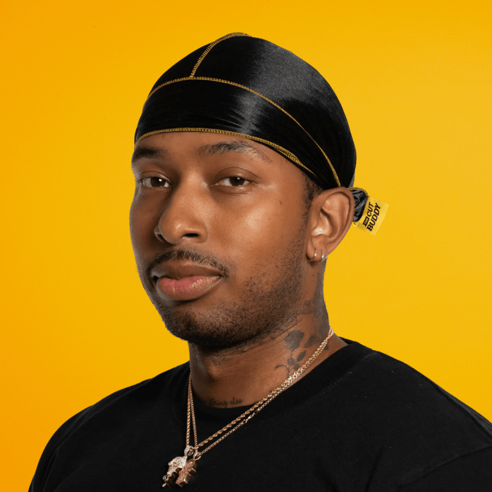 Three-quarter angled view of a model wearing the Nick Wavy black silky durag with yellow stitching against a yellow background.